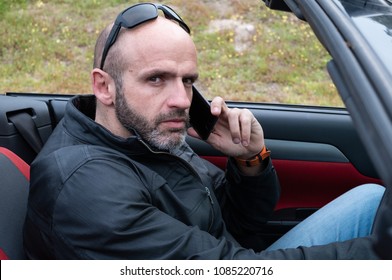 Middle-age Bald And Serious Man With Sunglasses And Leather Jacket Talking On His Smart Phone In His Convertible Car