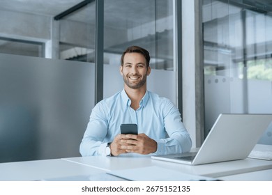 Middle-age manager man ceo using cell phone mobile app, laptop. Smiling Latin young businessman holding smartphone in business office working crypto online, looking at camera portrait. Copy space