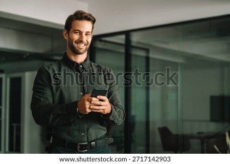 Middle-age manager man ceo using cell phone mobile application. Smiling Latin young businessman holding smartphone in business office working crypto online, looking at camera portrait. Copy space