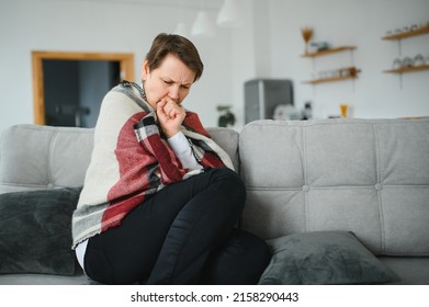 Middle-aged 50s sick frozen woman seated on sofa in living room covered with warm plaid sneezing, runny nose feels unhealthy, seasonal cold, weakened immune system concept - Shutterstock ID 2158290443