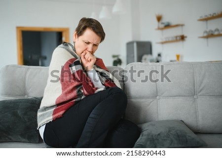 Middle-aged 50s sick frozen woman seated on sofa in living room covered with warm plaid sneezing, runny nose feels unhealthy, seasonal cold, weakened immune system concept