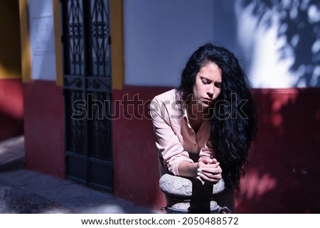 Middle-aged adult Hispanic woman with black curly hair, wearing a salmon-colored suit leaning on a column, sad and depressed. Concept sadness, depression, loneliness, grief.