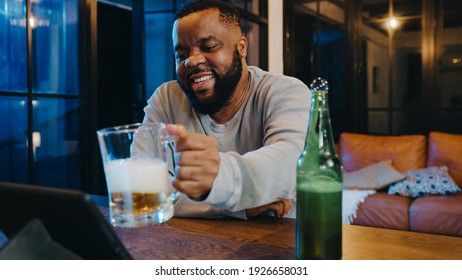 Middle-aged African American Male Drinking Beer Having Fun Happy Night Party Event Online Celebration Via Video Call In Living Room At Home. Social Distancing, Quarantine For Coronavirus Prevention.