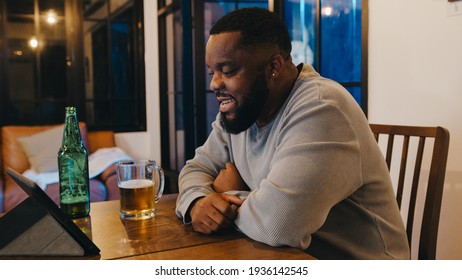 Middle-aged African American Male Drinking Beer Having Fun Happy Night Party Event Online Celebration Via Video Call In Living Room At Home. Social Distancing, Quarantine For Coronavirus Prevention.