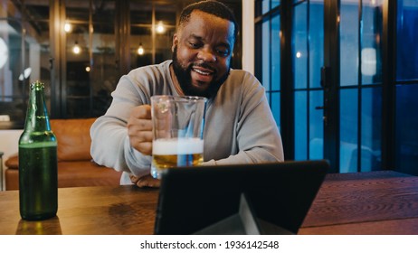 Middle-aged African American Male Drinking Beer Having Fun Happy Night Party Event Online Celebration Via Video Call In Living Room At Home. Social Distancing, Quarantine For Coronavirus Prevention.