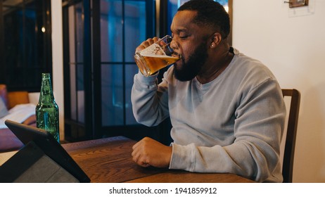 Middle-aged African American Male Drinking Beer Having Fun Happy Night Party Event Online Celebration Via Video Call In Living Room At Home. Social Distancing, Quarantine For Coronavirus Prevention.