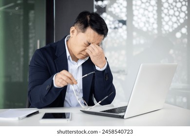 A middle-aged Asian businessman feeling overwhelmed and stressed in a modern office setting, massaging the bridge of his nose while looking at his laptop. - Powered by Shutterstock