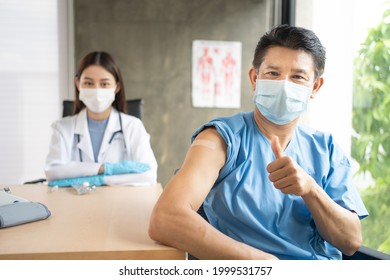 Middle-aged Asian man in a patient gown and face mask sitting at desk smiling showing thumbs up after got COVID-19 vaccination at arm with a blurred Asian female doctor in face mask sit in the back. - Powered by Shutterstock