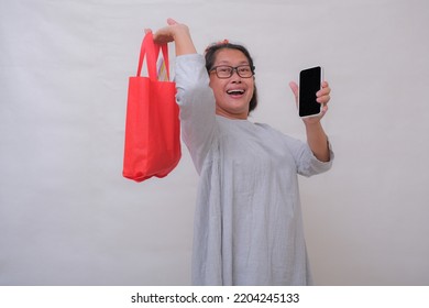 A middle-aged Asian woman holds a red shopping bag in one hand, a cell phone in the other; smiling, cheerful, happy expression. - Shutterstock ID 2204245133