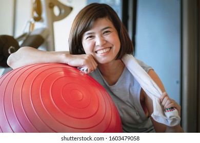 Middle-aged Asian Woman Smiling Brightly While Sitting At The Red Yoga Ball In The Gym