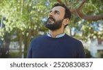 Middle-aged bearded man in contemplation amidst green park foliage, exuding serenity and introspection.