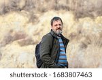 A middle-aged bearded man with a dark backpack stands on the shore of a flooded quarry.