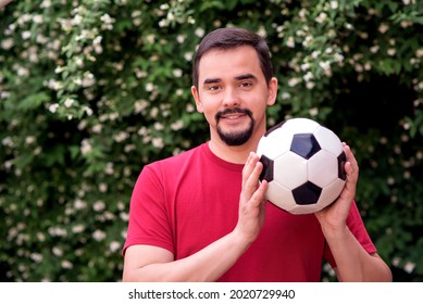 Middle-aged Bearded Man In Red Shirt Holding Football Ball And Smiling: White Jasmine Flowers And Green Leaves Behind. Happy Active Man Dad Or Coach With Soccer Ball In Arms. Healthy Lifestyle Concept