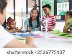 A middle-aged biracial teacher and four young students craft together in a classroom. They are surrounded by colorful papers and sustainability posters, learning, education, unaltered