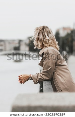 A middle-aged blonde woman with curly hair stands on the river embankment in the city