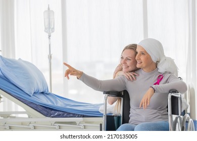 A Middle-aged Breast Cancer Woman With Clothing Around Her Head Affected From Chemotherapy Sitting On Wheel Chair And Pointing Finger To Somewhere With Her Daughter.