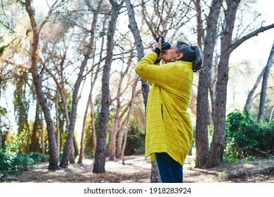 Middle-aged Brunette Woman With Gray Hair And Yellow Jacket, Takes Photos With Analog Reflex Camera In The Forest