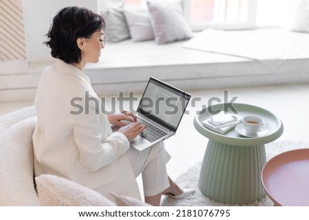 Middle-aged business woman is typing in a word document on a laptop while sitting on a couch in her home office. Distant work. 