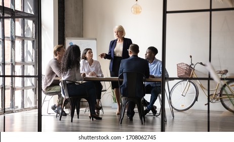 Middle-aged businesswoman lead meeting with multicultural businesspeople in office brainstorm together, confident mature female boss speak talk at briefing with employees, leadership concept