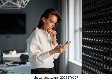 A middle-aged businesswoman standing in the office and using a smartphone. Attractive female looking thoughful and text messaging. 