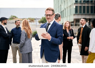 A middle-aged Caucasian business man worker using a tablet in formal attire, standing in front of a crowd of people at a public event gesturing and typing digital display information - Powered by Shutterstock