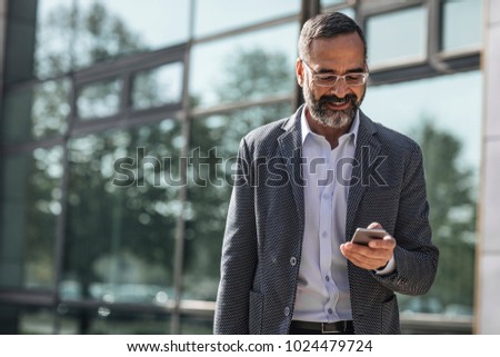 Middle-aged Caucasian smiling businessman standing outdoors and reading on his cell phone.