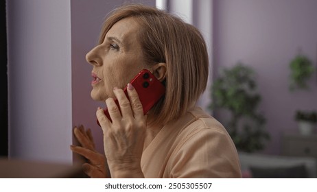 A middle-aged caucasian woman with blonde hair is indoors at home, holding a red phone and looking out through a window in a cozy living room setting. - Powered by Shutterstock