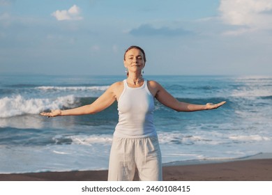 A middle-aged caucasian woman practices qigong energy exercise outdoor on the beach, blue sky and ocean backdrop, charges life force energy, esoteric practitioner, energy healer, grounding healing - Powered by Shutterstock