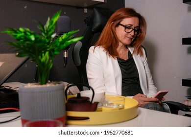 Middle-aged Caucasian Woman Sitting In A Chair Next To A Desk At Home After Taking An Infusion Looking At The Mobile Phone