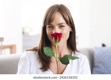 A middle-aged Caucasian woman smiles while holding a red rose close to her face. Her joyful expression suggests a moment of happiness or a romantic gesture. - Powered by Shutterstock