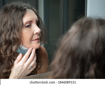 Middle-aged caucasian woman suffering with acne on her chin after wearing protective face mask. Looking at mirror. 