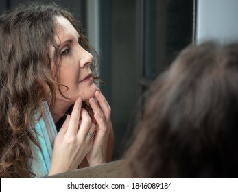 Middle-aged Caucasian Woman Suffering With Acne On Her Chin After Wearing Protective Face Mask. Looking At Mirror. 