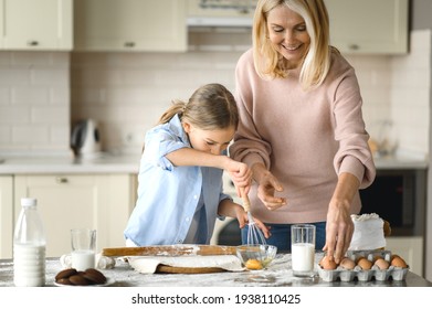 A middle-aged caucasian woman teaches a little granddaughter to cook. Grandma teaches her granddaughter to beat eggs, they make and roll the dough, use ingredients, cooking together