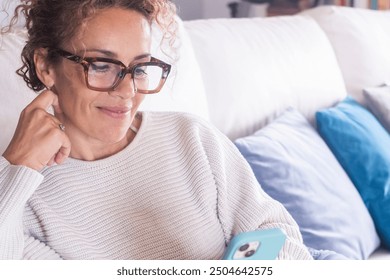 Middle-aged caucasian woman wearing reading glasses, comfortably seated on her home couch, smiling while using her smartphone to browse social media, download, use apps, reflecting a modern lifestyle - Powered by Shutterstock