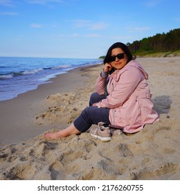 A Middle-aged Chubby Female In A Pink Rainjacket Sitting And Relaxing On The Beach With Her Shoes Off