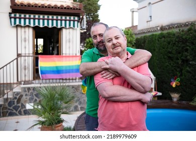 Middle-aged Couple Embracing In The Garden Of Their Home With An LGBT Flag In The Background.