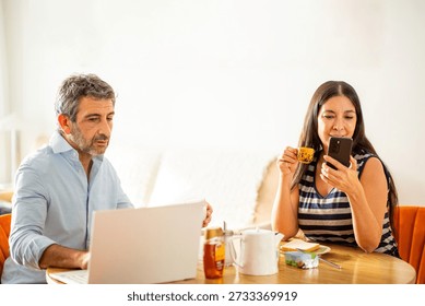 Middle-aged couple having breakfast at the table, with one using a laptop and the other checking her phone while sipping coffee