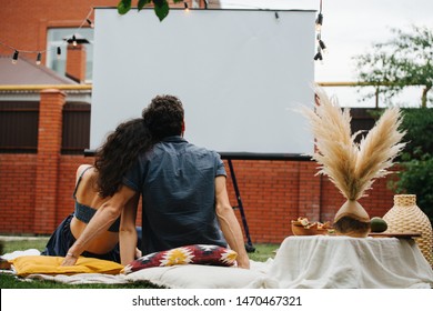 Middle-aged couple in love is watching a movie, leaning to each other during a picnic on the lawn in their courtyard, behind a brick fence. Blank empty white screen.