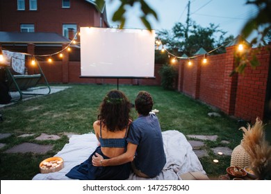 A middle-aged couple in love is watching a movie, in the twilight, outside on the lawn in their courtyard. They are sitting on a tablecloth, watching a projector screen. Man is hugging his wife.