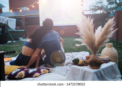 A Middle-aged Couple In Love Is Watching A Movie, In The Twilight, Outside On The Lawn In Their Courtyard. They Are Sitting On A Tablecloth, Watching A Projector Screen, Leaning To Each Other.