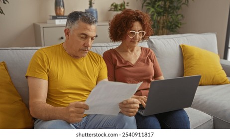 Middle-aged couple sitting together on a couch at home, with a man reviewing documents and a woman using a laptop in the living room setting, reflecting a domestic and focused atmosphere - Powered by Shutterstock