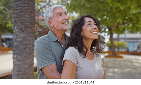 Middle-aged couple smiling happily in a sunny urban park setting, with man embracing woman amidst lush greenery, capturing the essence of love and togetherness outdoors. - Powered by Shutterstock