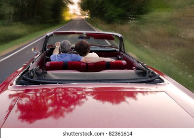 A Middle-aged Couple Snuggle Together As They Zoom Down A County Road In A Classic Red Convertible, Perhaps On A Sunday Drive As They Enjoy The Experience And Each Others Company.