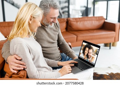 Middle-aged couple using laptop for video connection with grown children, grey-haired man and blonde woman talks with couple of young people on the screen. Video chatting concept, side view - Powered by Shutterstock