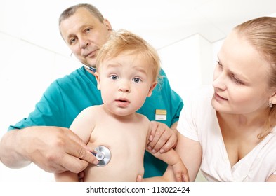 A Middle-aged Doctor In A Green Smock Is Listening To A Baby's Back With A Phonendoscope In A Medical Study. The Baby Is 1,5 Years Old. He Has A Surprised Look. Caucasian. Focus Is On The Boy.