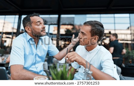 A middle-aged father gestures while having a serious or difficult conversation with his adolescent son at a restaurant terrace.