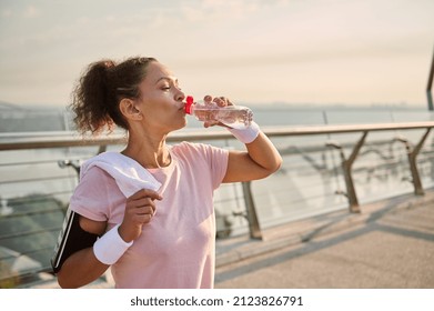 Middle-aged female athlete in a pink T-shirt and gray leggings, with white wristbands, holds a terry towel on her shoulders and drinks water after workout standing on the city bridge at sunrise