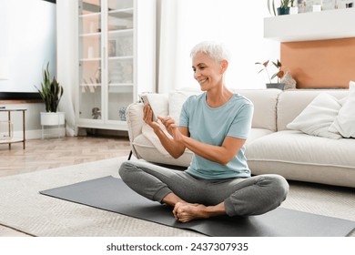 Middle-aged flexible woman in fitness outfit sitting in lotus position while using cellphone for sport app. Caucasian mature woman doing online training tutorial - Powered by Shutterstock