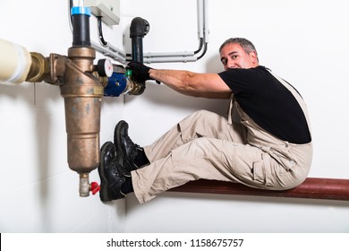 Middle-aged Funny Grey Haired Plumber In Black T-shirt, Dirty Coveralls, Textile Gloves And Boots Repairing Or Breaking Metallic Water Pipes With Hands' Power On The Background Of White Wall