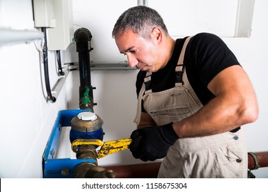 Middle-aged grey haired plumber in black t-shirt, dirty coveralls and black textile gloves repairing metallic water pipes with yellow wrench near electrical boards on the background of white wall